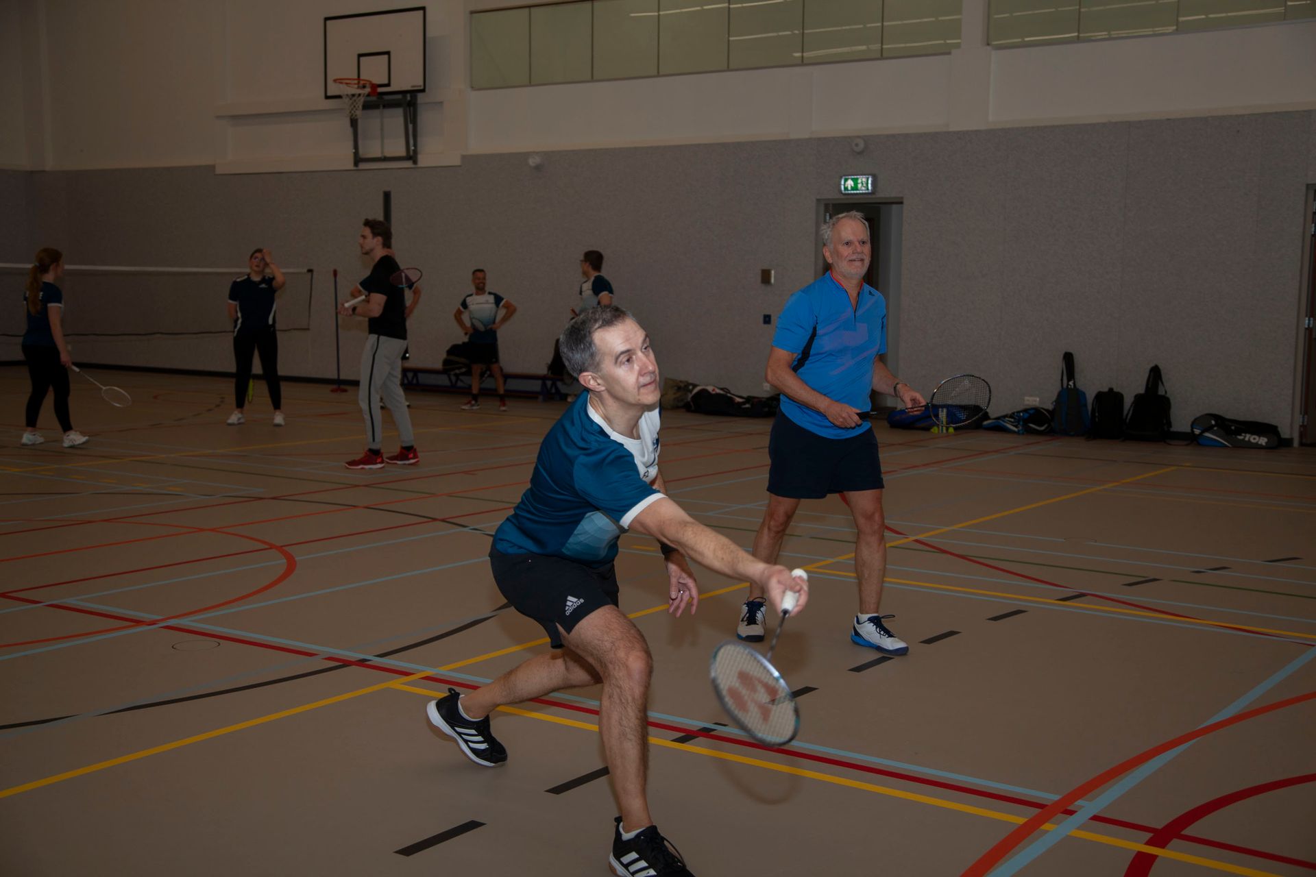 Mensen spelen badminton in een sporthal, waarbij één persoon op de voorgrond probeert de shuttle met een racket te raken.