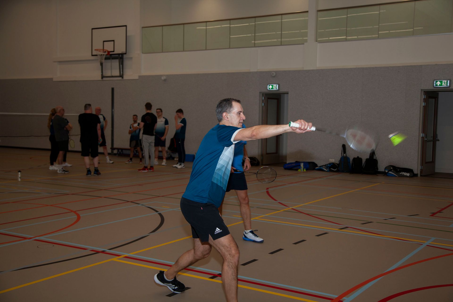 Een persoon in een blauw shirt zwaait met een badmintonracket naar een shuttlecock op een indoorbaan