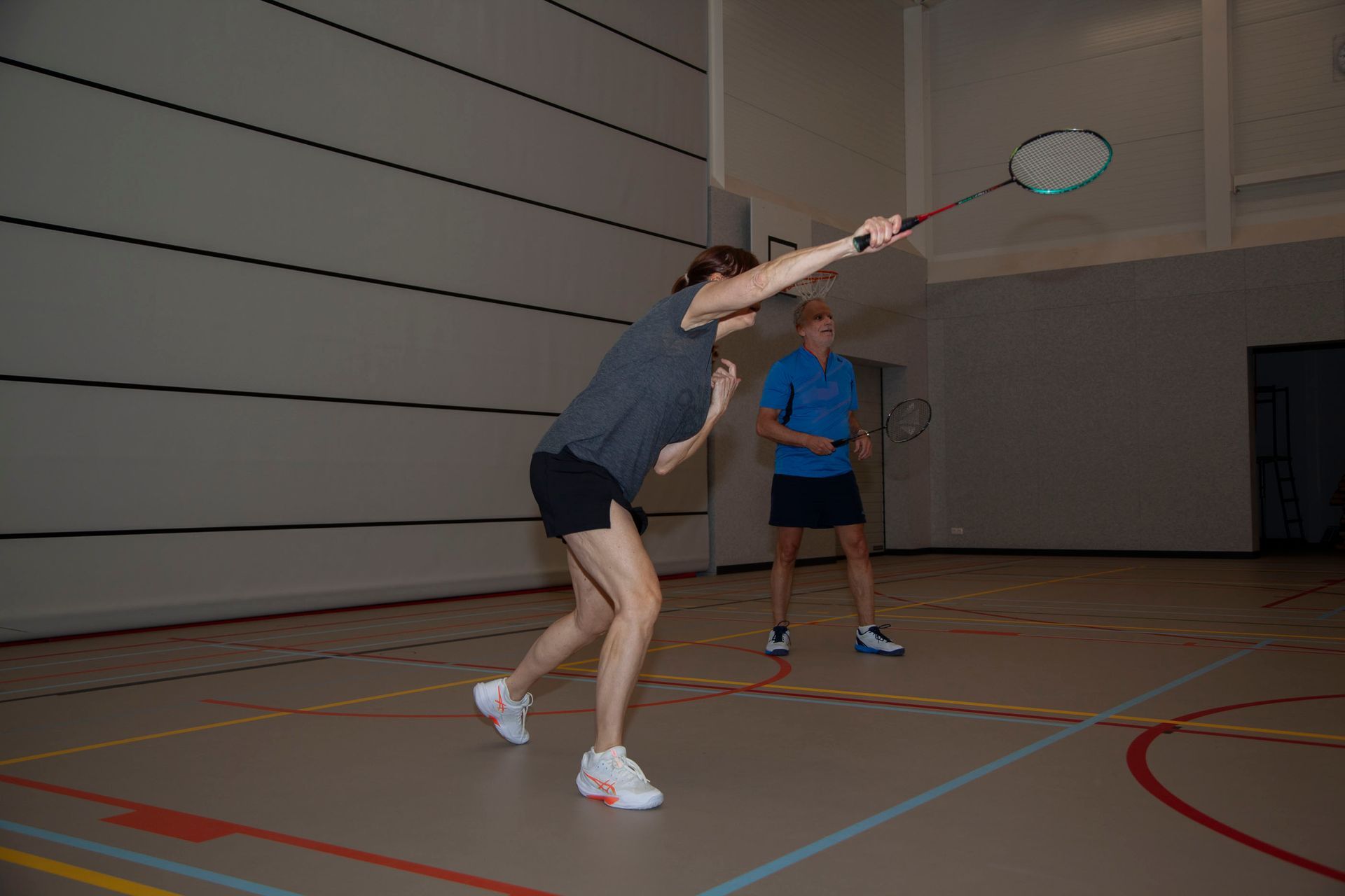 Een persoon in een grijs shirt slaat met een racket een shuttlecock in een overdekte badmintonhal