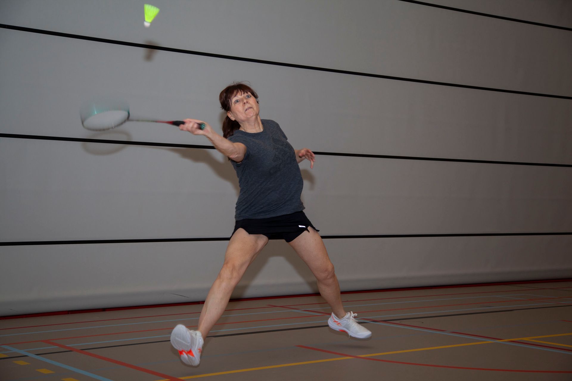 Een persoon in een grijs shirt en een donkere korte broek slaat met een racket tegen een felgele badmintonshuttle