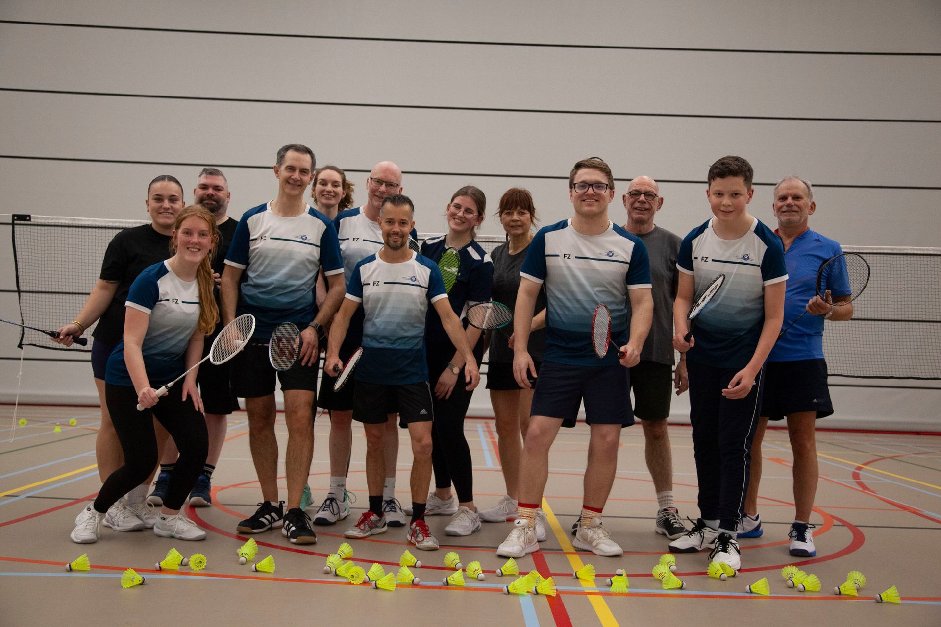 Een groep badmintonspelers in blauw-witte shirts poseert samen op een indoorbaan, omringd door shuttlecocks.