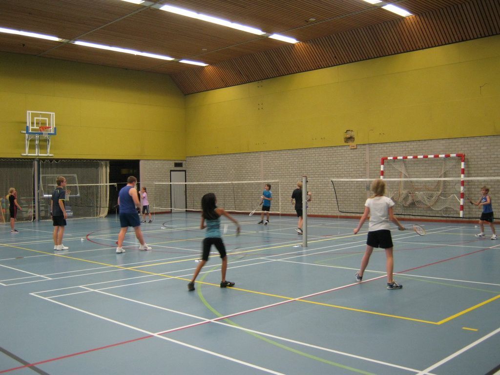 Mensen spelen badminton op een blauwe indoorbaan met gele muren onder fel licht.