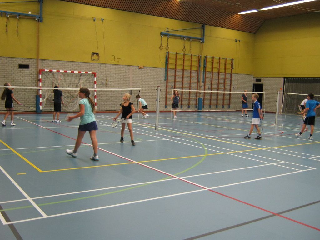 Mensen spelen badminton op indoorbanen in een sporthal met gele muren en houten plafondpanelen.