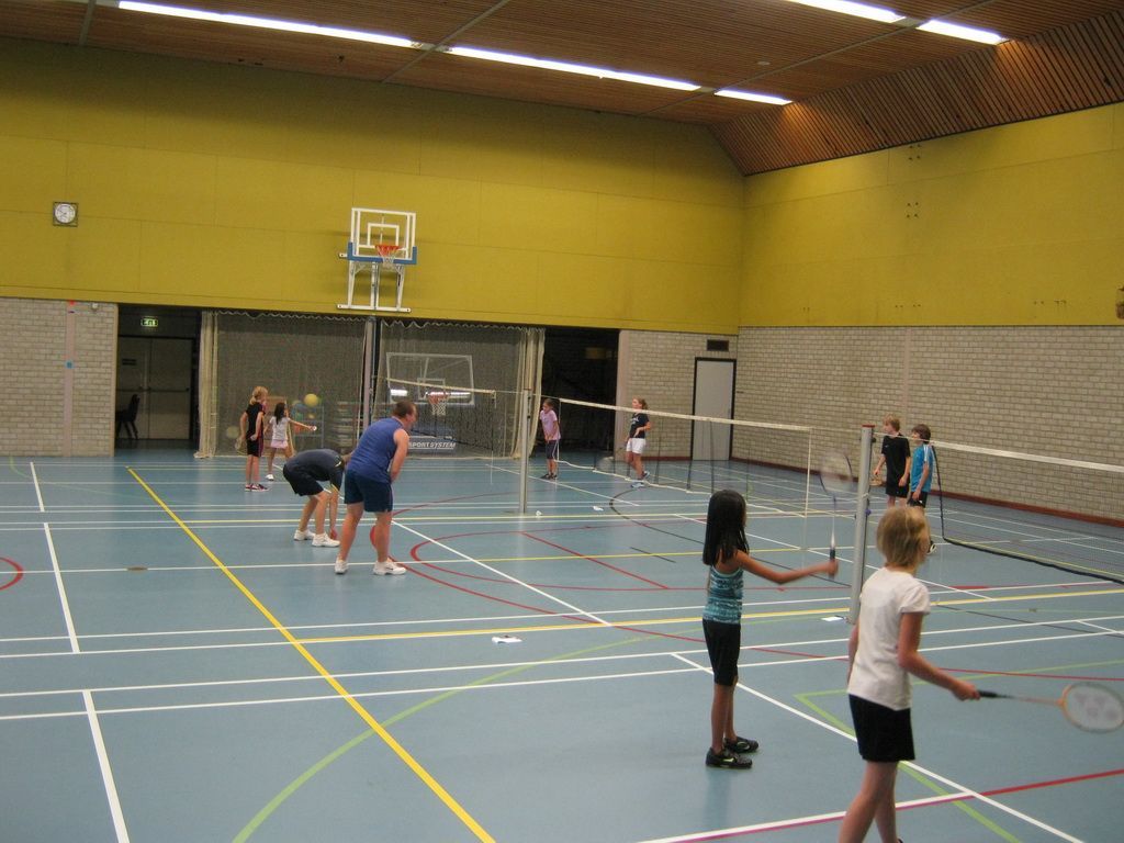 Mensen spelen badminton in een sporthal met gele muren, gemarkeerde vloeren en een blauwe baan.
