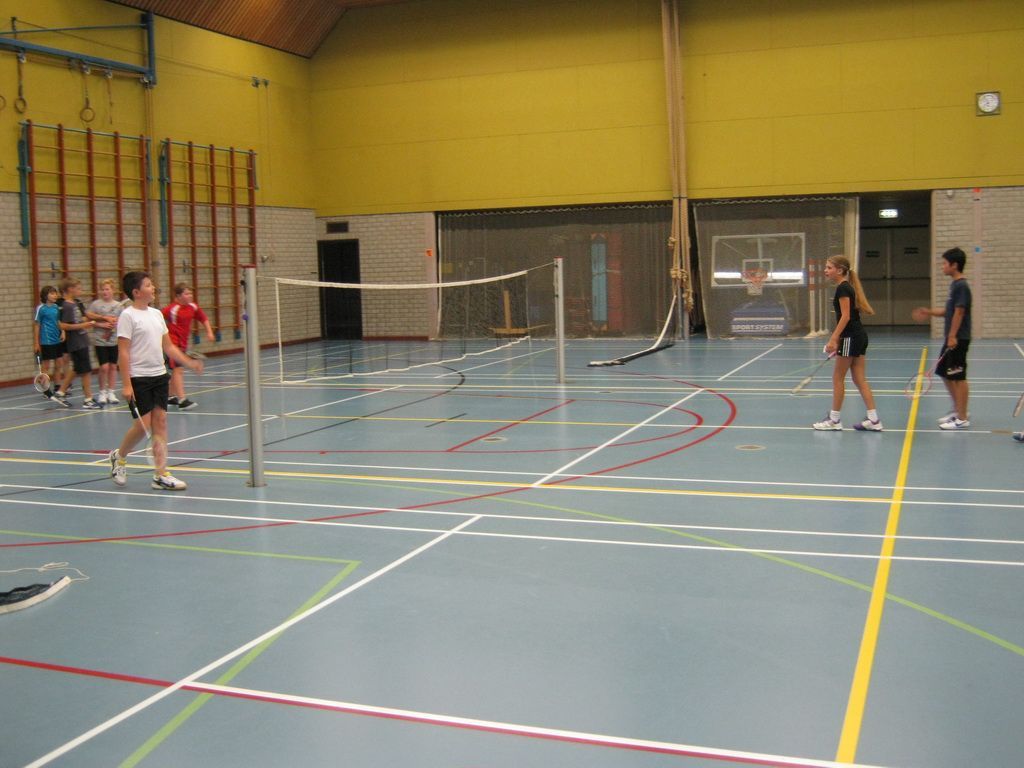 Een groep mensen speelt badminton in een overdekte sporthal met gele muren, meerdere netten en veldlijnen op de vloer.
