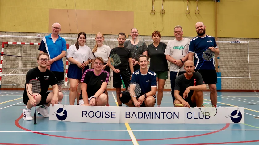 Een groep mensen met badmintonrackets poseert achter een bord van de Rooise Badminton Club op een indoor sportveld.