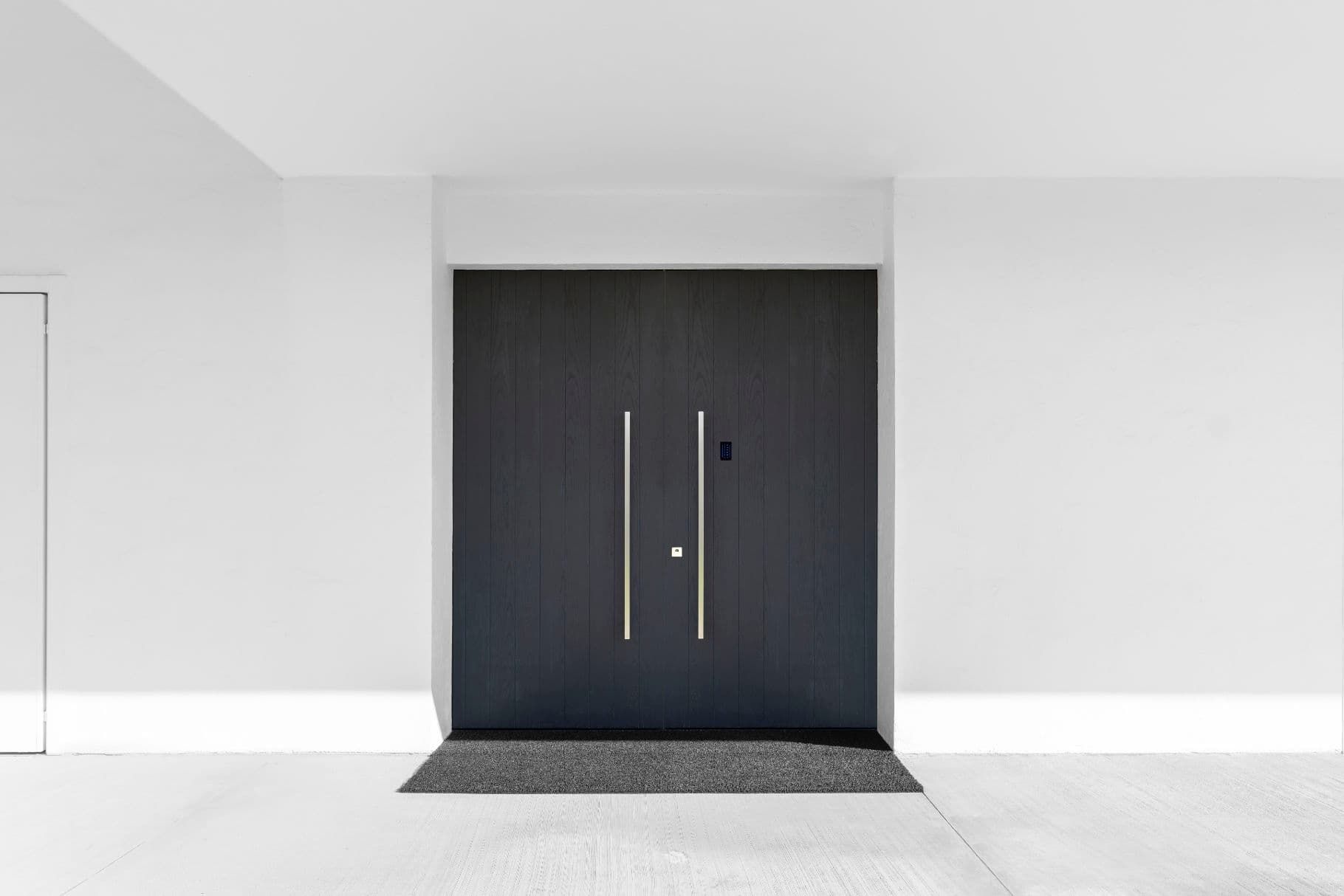 Porta d'ingresso grigio scuro con due maniglie verticali argentate, zerbino nero, pareti bianche.