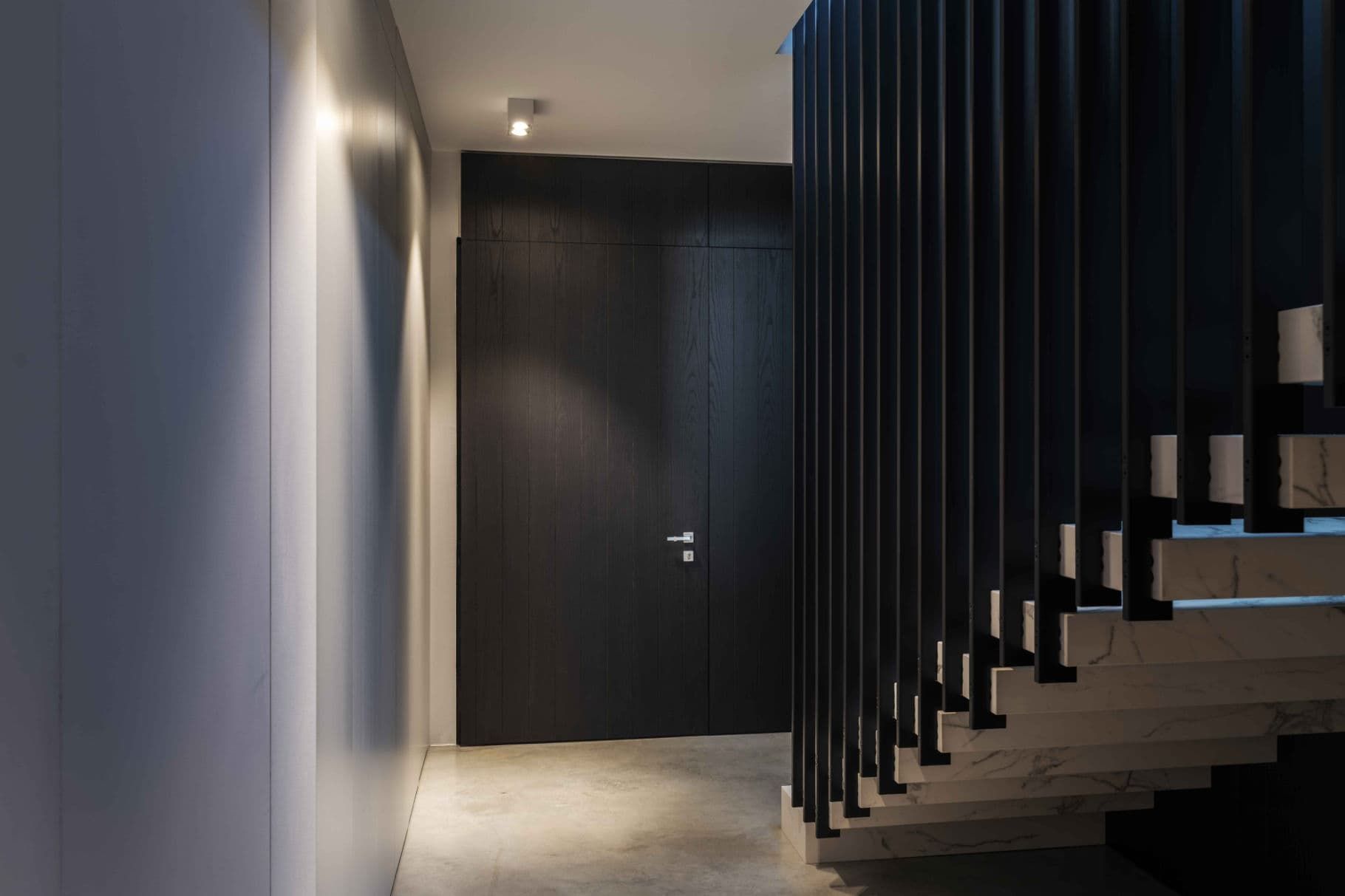 Corridoio con porta in legno scuro e scala con ringhiera nera. Pavimento in cemento, pareti bianche.