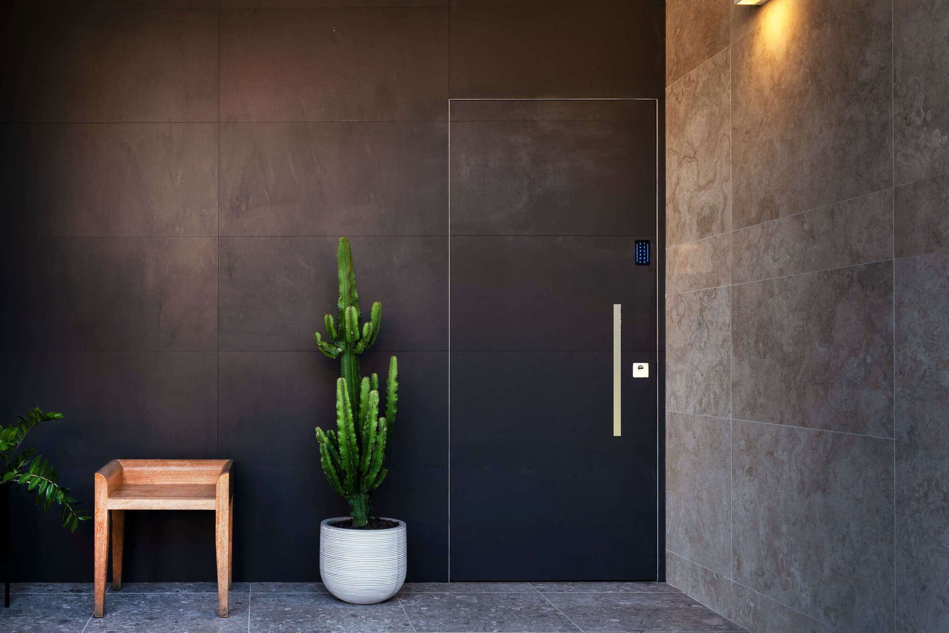 Moderna porta nera con maniglia verticale dorata, cactus, piccola sedia di legno e pareti in pietra.