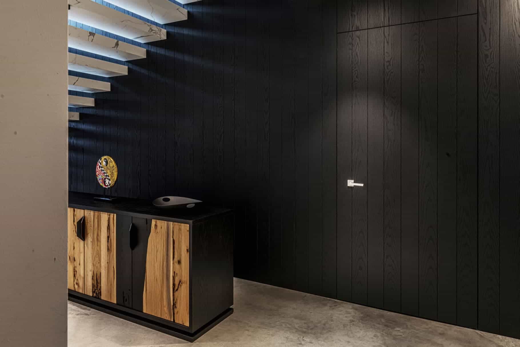 Parete rivestita in legno nero con porta nascosta. Mobiletto in legno con scultura dorata in cima.