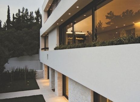 Moderno edificio bianco con grandi finestre che riflettono la luce interna, accanto a un cortile alberato.