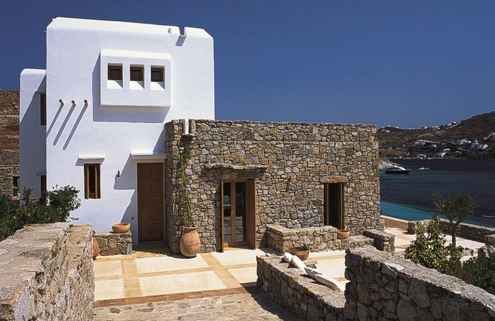 Villa bianca e in pietra in riva al mare, cielo azzurro. Patio terrazzato con muri in pietra, porta marrone e pianta in vaso.
