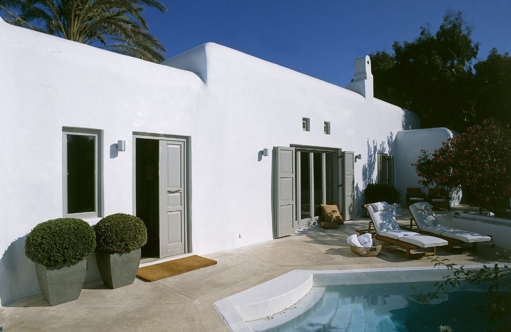 Casa in stucco bianco con porte aperte e piscina. Due cespugli in vaso sul davanti, sedie a sdraio a bordo piscina.