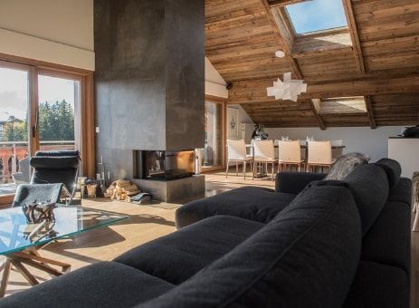 Accogliente soggiorno con camino, soffitto in legno, divano scuro, tavolo in vetro e zona pranzo visibile sullo sfondo.