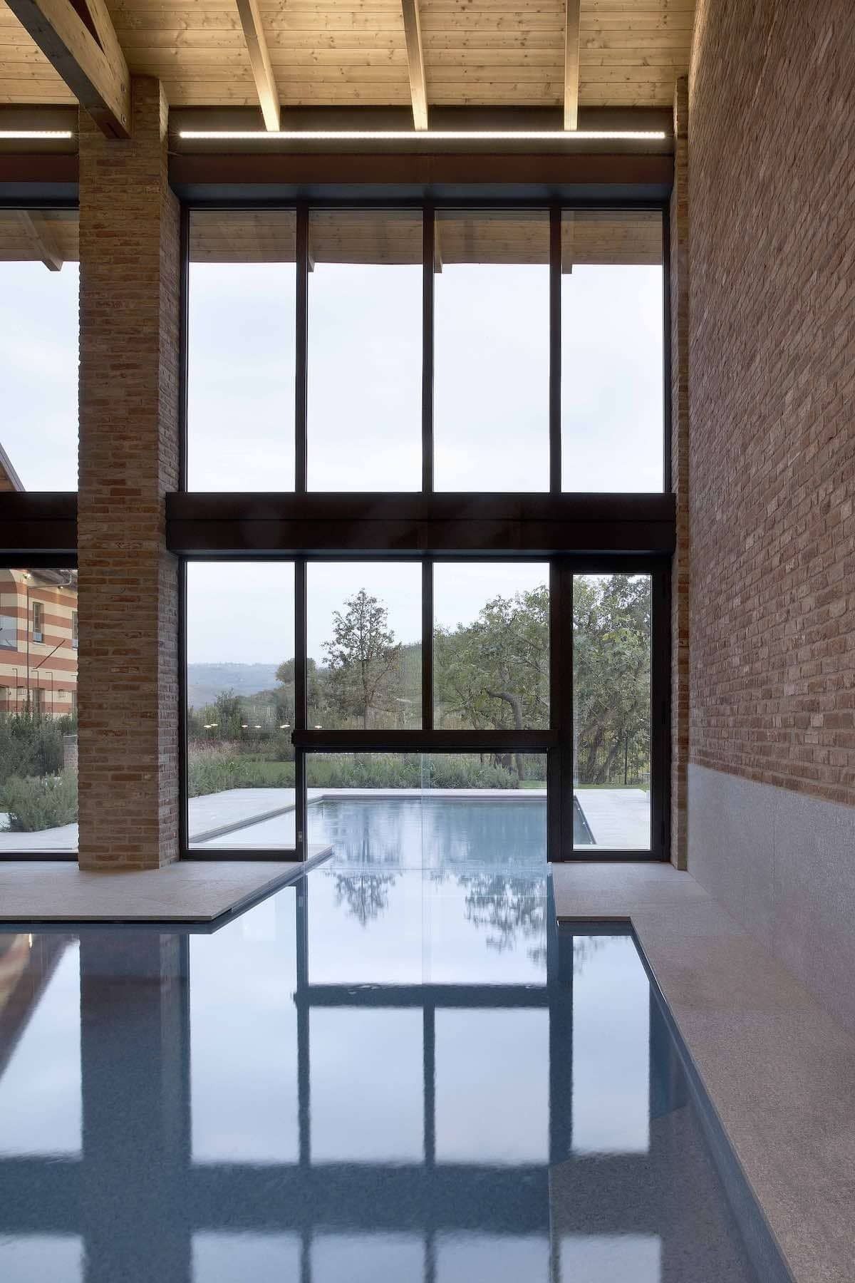 Piscina coperta con ampie vetrate che si affacciano sugli alberi. Pareti in mattoni e travi in legno incorniciano la vista.