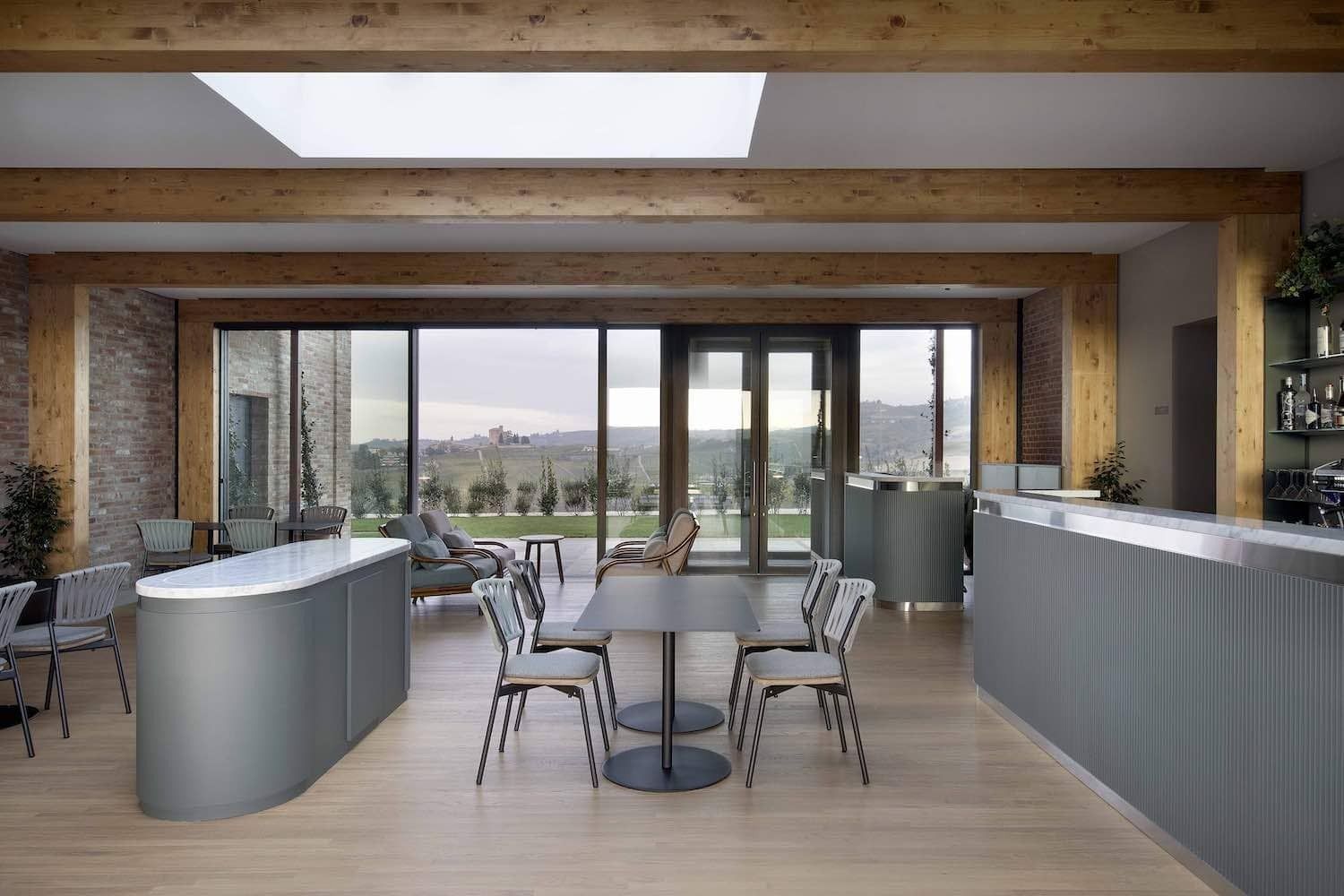 Interno di un bar moderno con ampie finestre, travi in legno, mobili grigi e un tavolo con sedie.