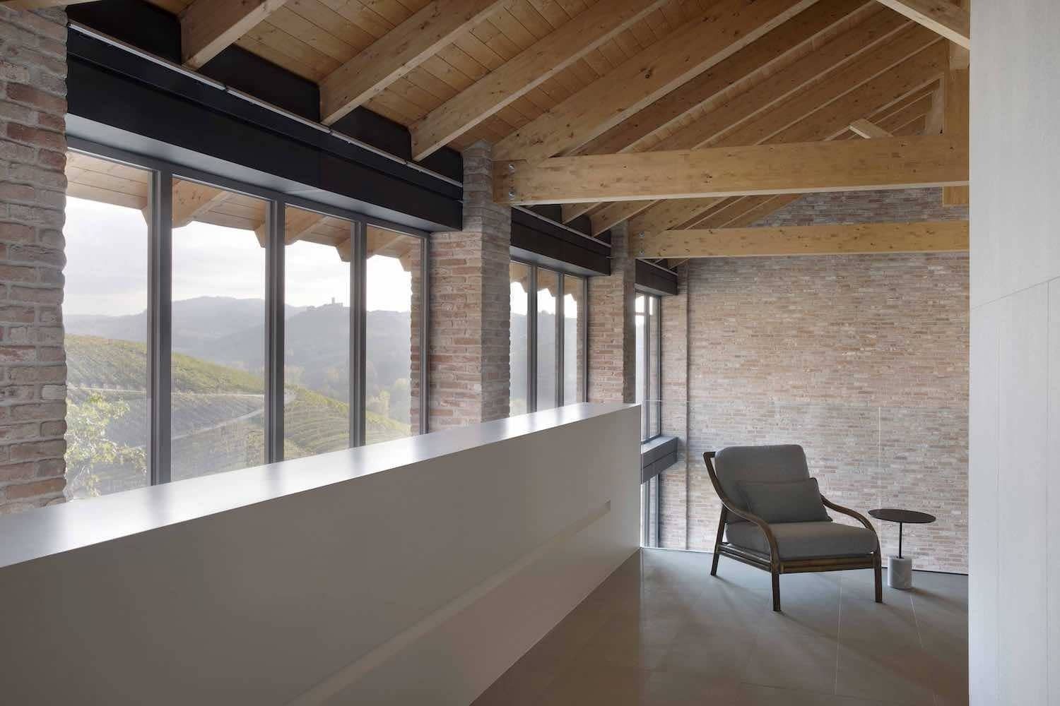 Interno con pareti in mattoni, travi in legno, grandi finestre, ringhiera bianca e una sedia.