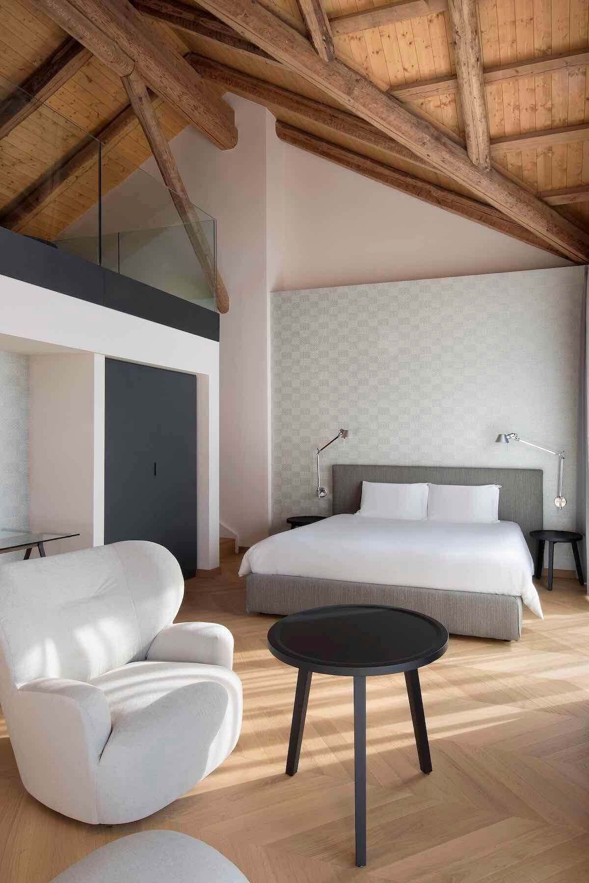 Camera da letto con travi in legno a vista, letto, sedia bianca e tavolino nero.