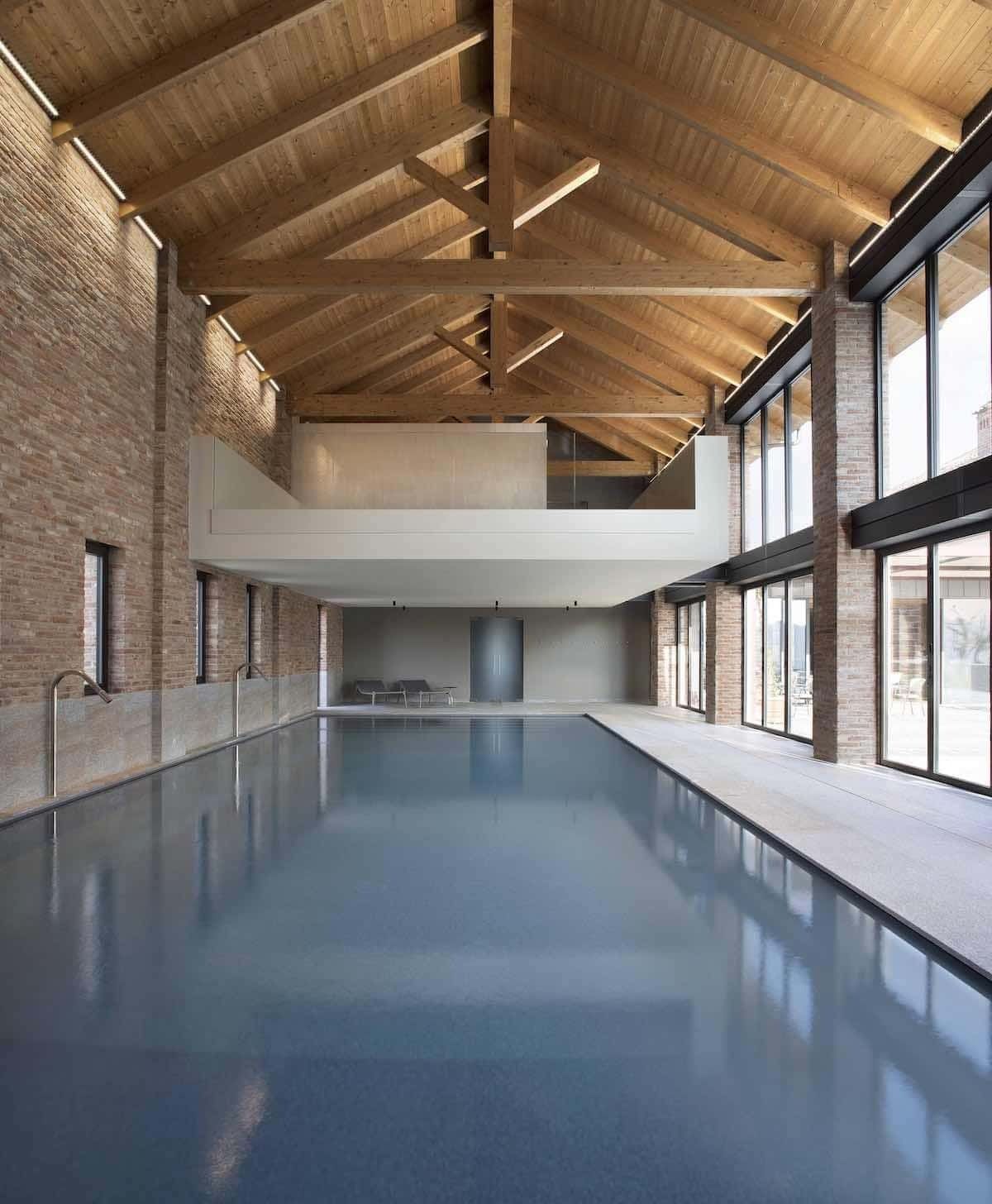 Piscina coperta con travi in legno a vista, pareti in mattoni e ampie finestre.