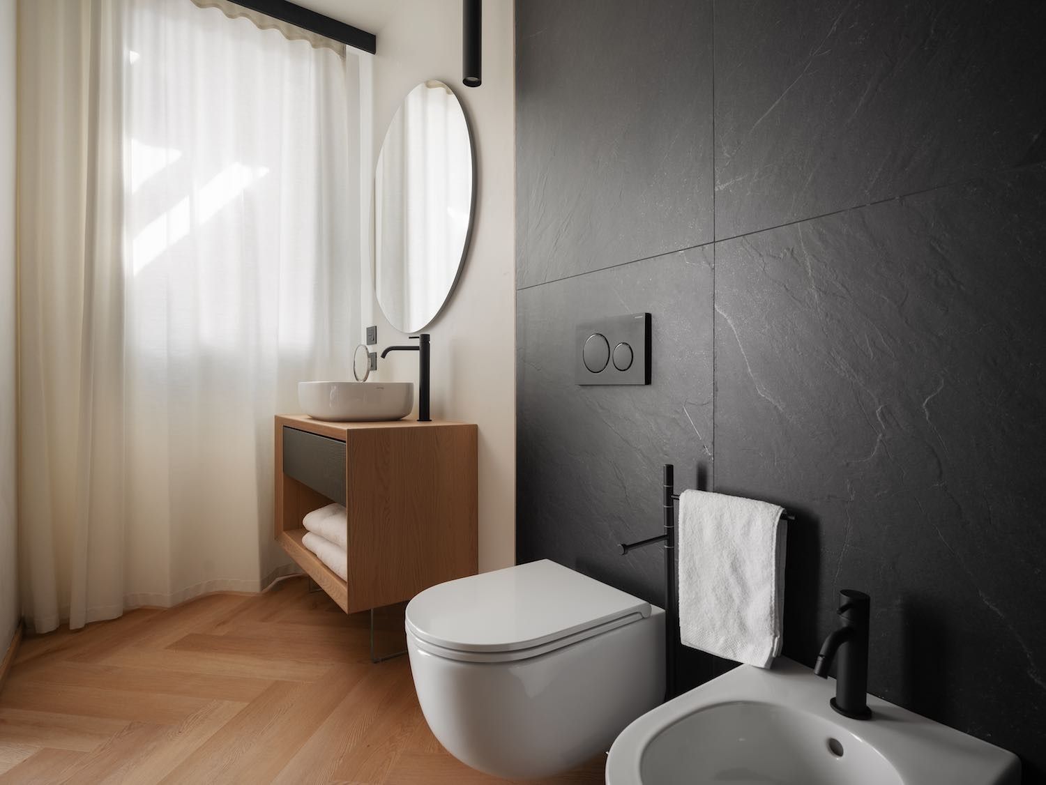Bagno con parete piastrellata nera, mobile lavabo in legno sospeso, WC bianco e bidet.