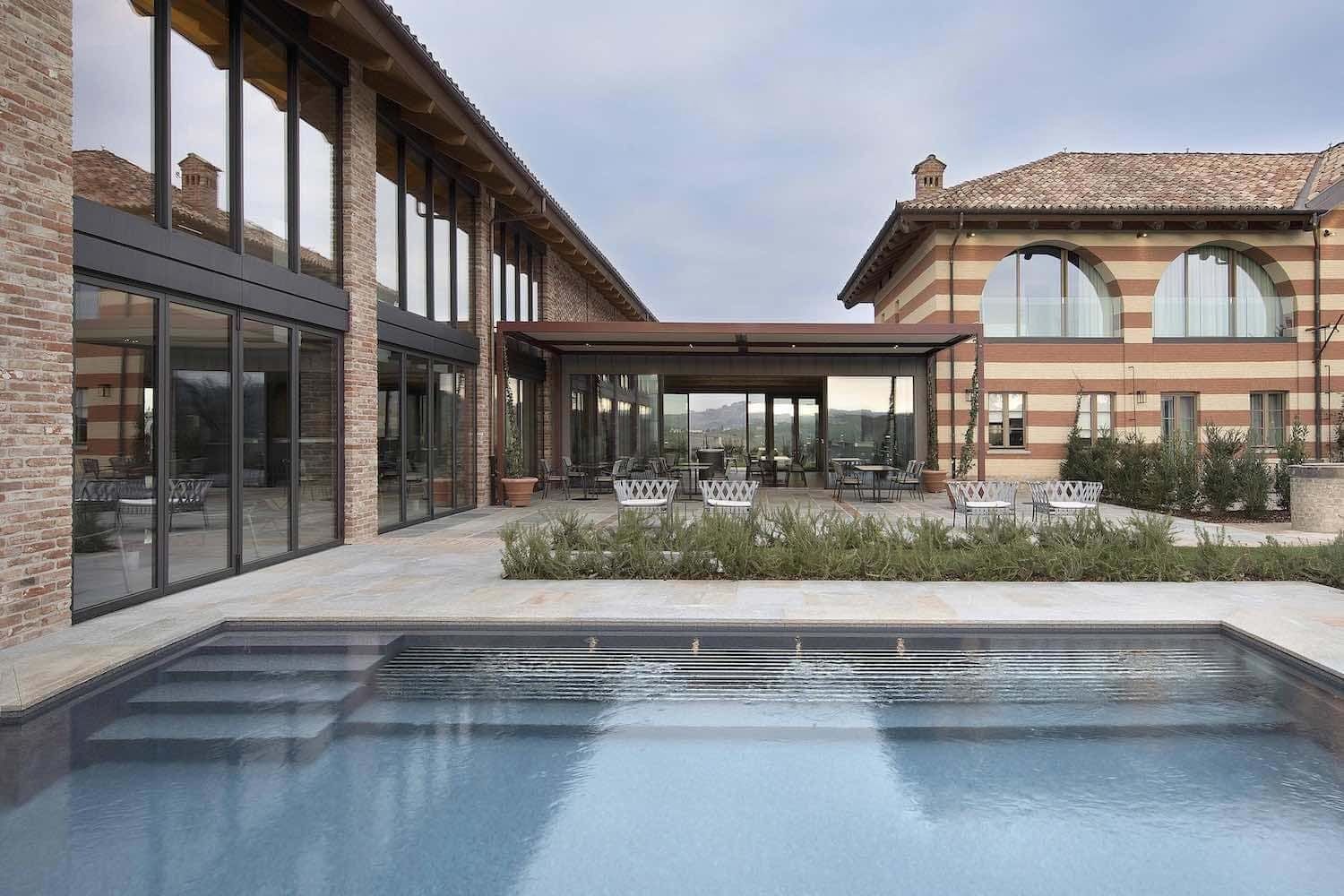 Piscina di fronte a un edificio moderno con ampie finestre e area salotto esterna coperta.