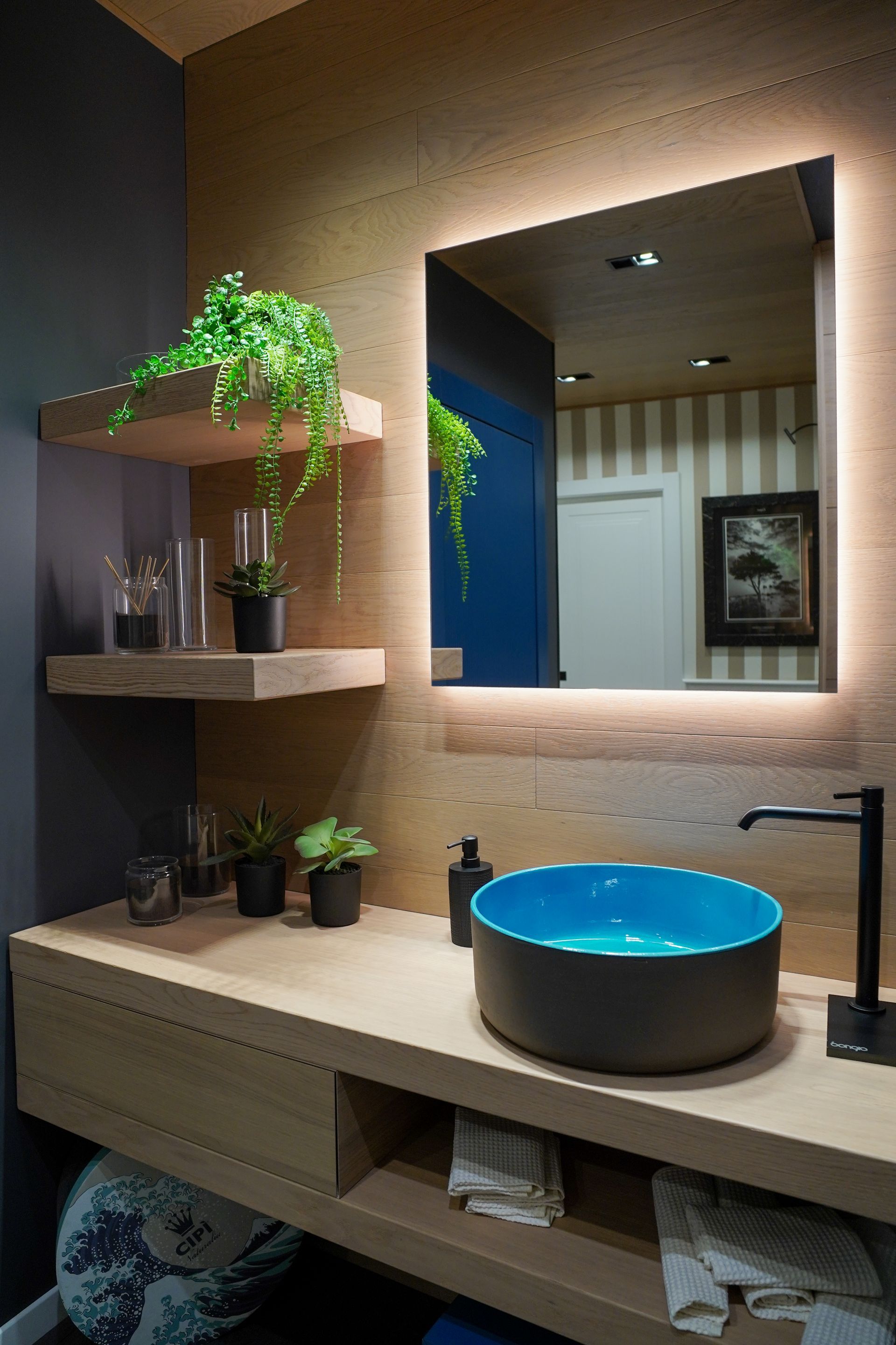 Bagno moderno con mobiletto color legno, lavandino blu, rubinetto nero e specchio retroilluminato.