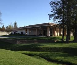 Low, beige building with a flat roof on a grassy lawn, framed by tall trees in bright daylight