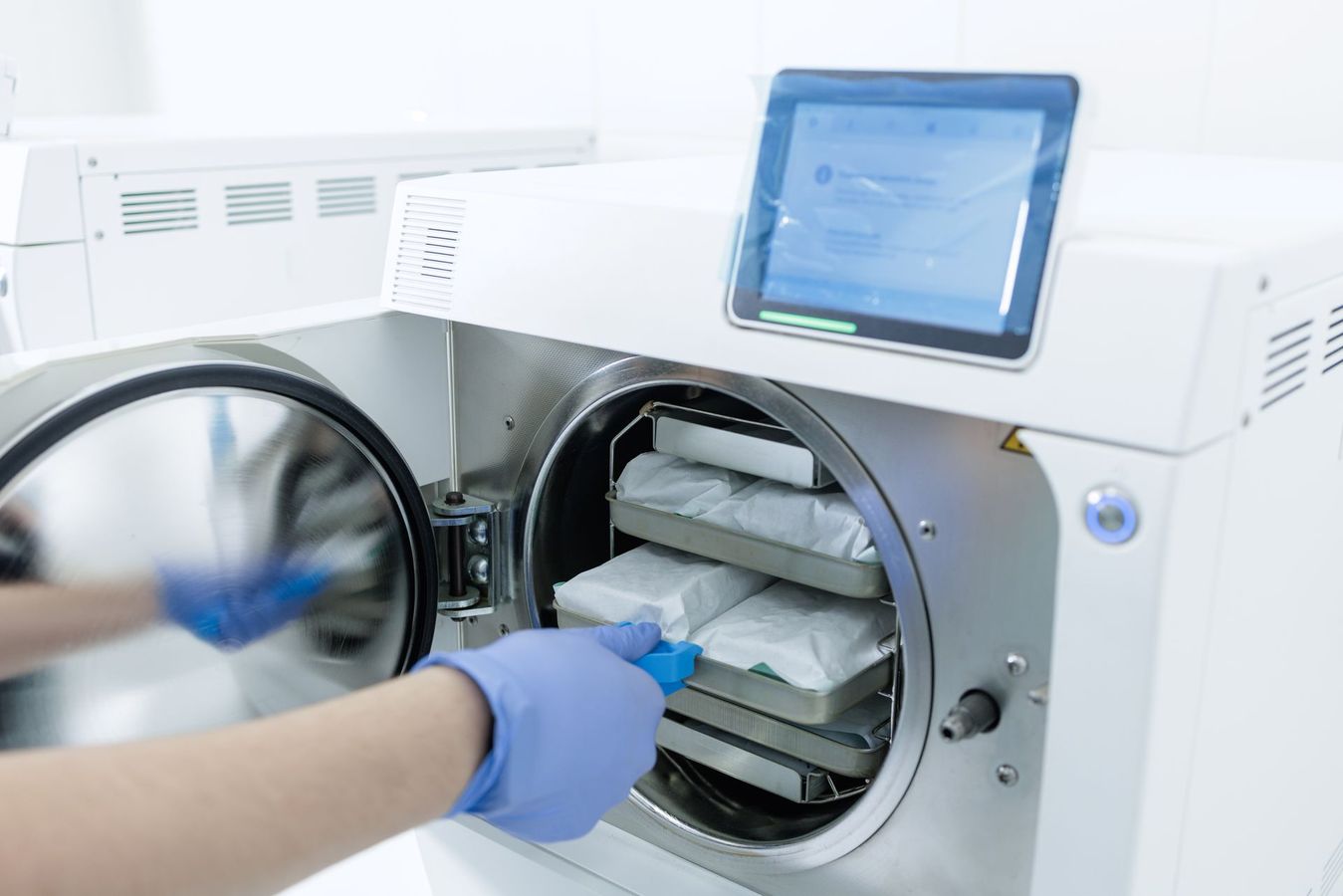 Gloved hands loading instruments into a sterilizer with an open round door and touchscreen display