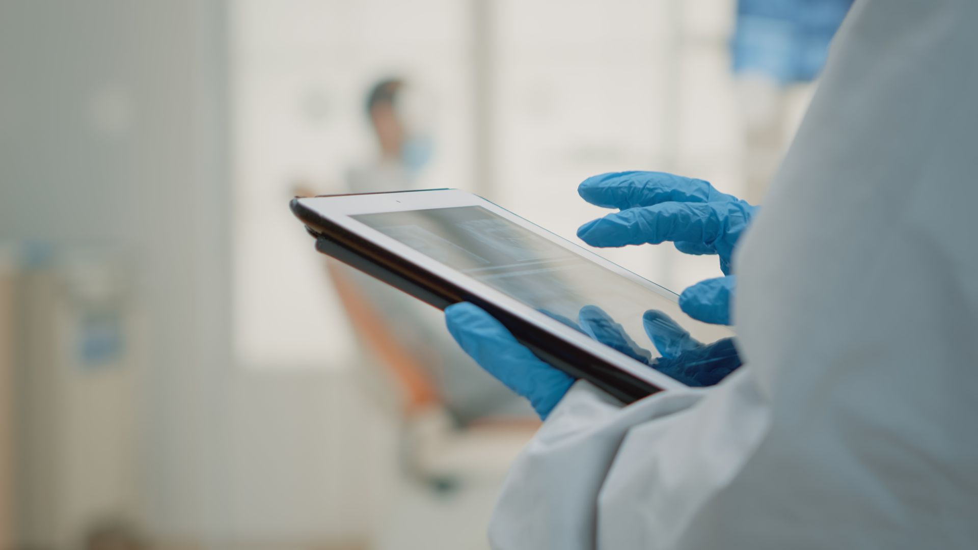 Gloved hands using a tablet in a clinical setting with a blurred patient in the background