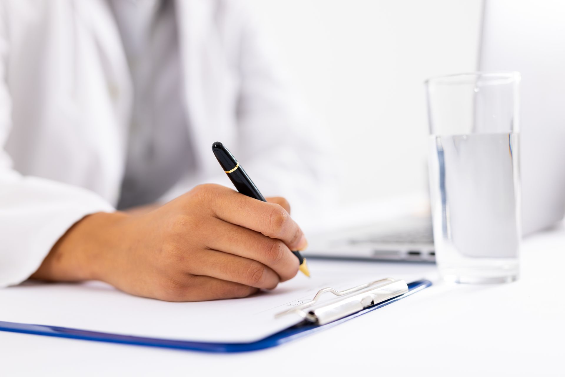Hand writing on a clipboard beside a glass of water on a white desk