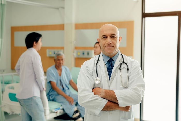 Doctor standing with arms crossed in a hospital room, with staff and a patient in the background