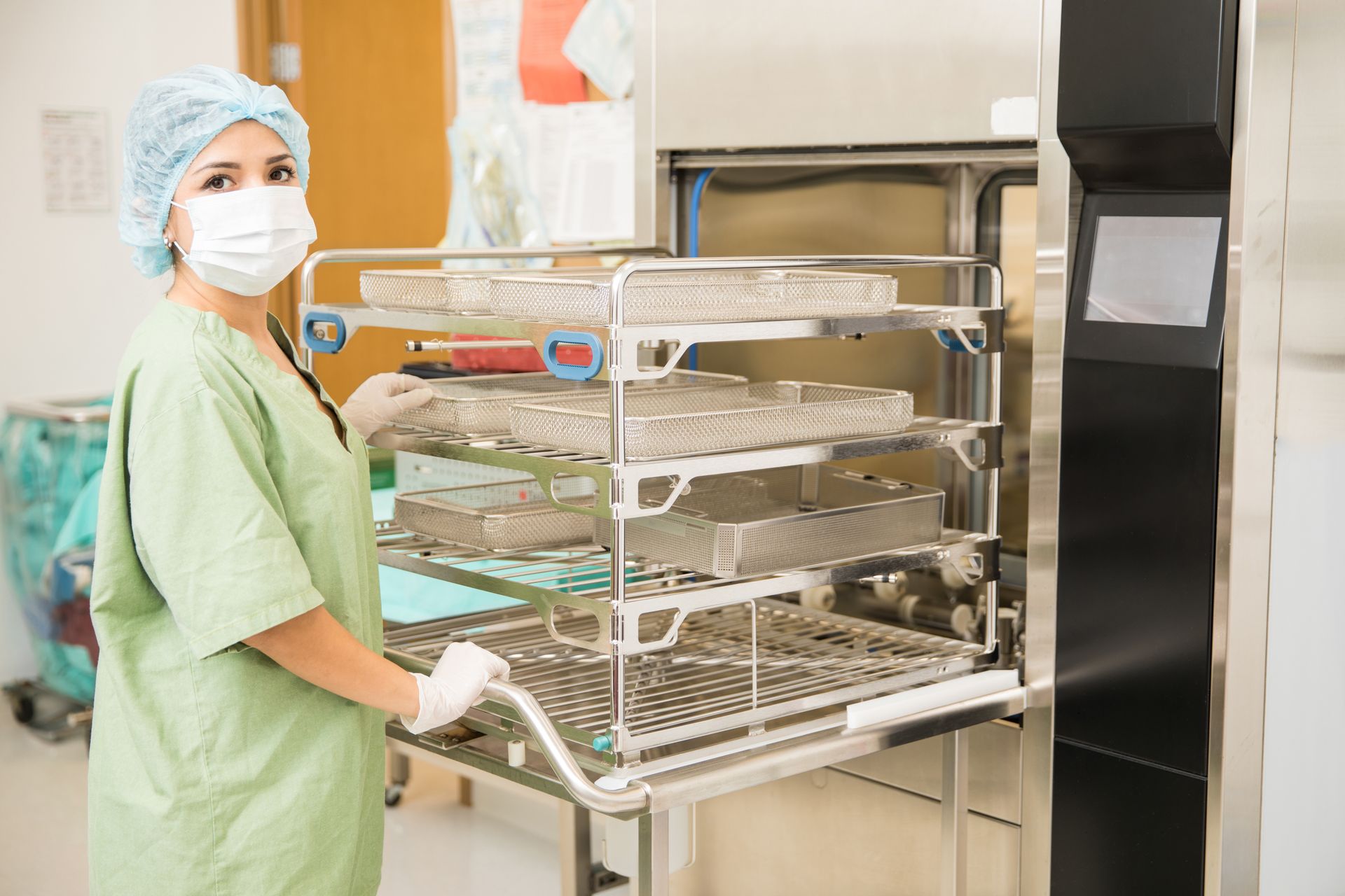 Hospital worker in green scrubs and mask handling a metal tray cart in a clinical kitchen or sterilization area