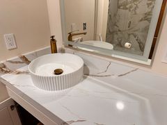 A white ribbed vessel sink sits on a white marble vanity with gold veining, featuring a gold faucet and a backlit mirror.