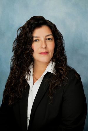 Studio portrait of a woman in a black blazer and white blouse against a blue-gray backdrop