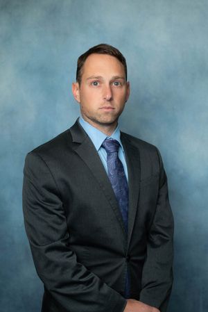 Formal portrait of a man in a dark suit and blue tie against a blue studio backdrop