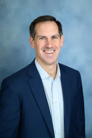 Smiling professional in a navy blazer and light blue shirt against a blue studio backdrop