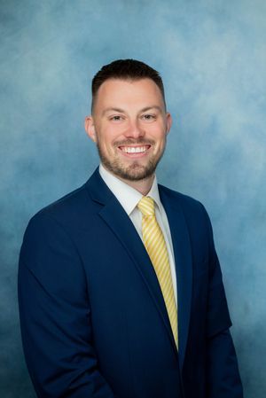Professional headshot of a smiling man in a blue suit, white shirt, and yellow tie against a blue backdrop