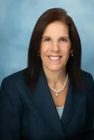 Professional headshot of a smiling woman in a dark blazer and pearl necklace against a blue background