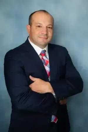 Man in a dark suit with a patterned tie, standing with arms crossed against a blue studio backdrop