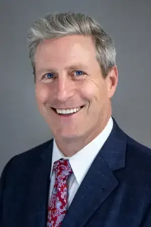 Professional headshot of a smiling man in a navy suit, white shirt, and red patterned tie on a gray background