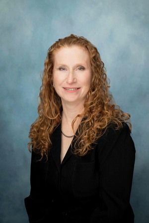 Studio headshot of a smiling person with curly red hair in a black blazer against a blue background