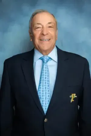 Smiling man in a dark suit with a light blue tie, posed against a blue studio backdrop.