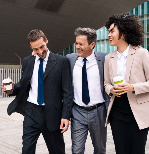 Three coworkers in business attire walking outdoors, smiling and holding coffee cups.