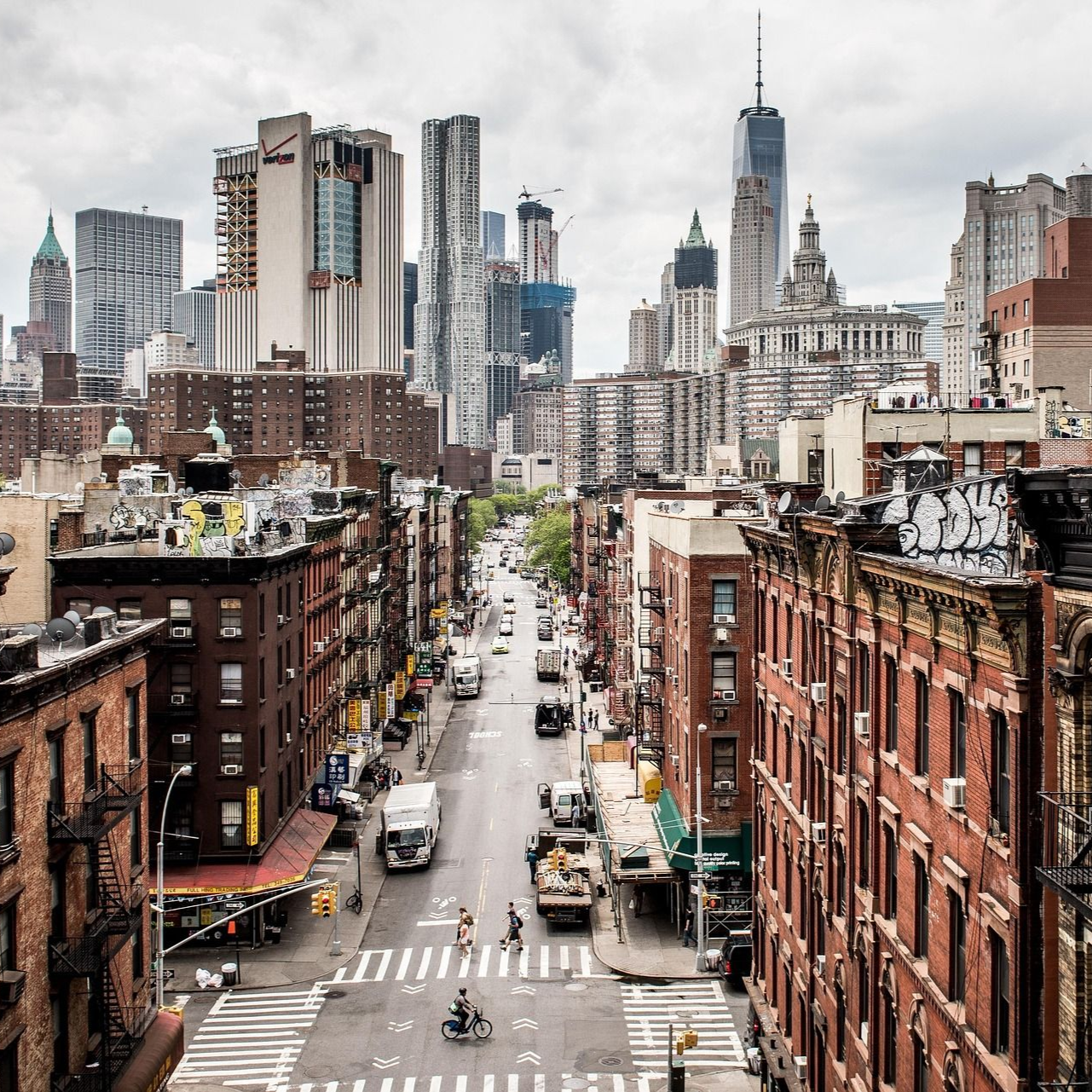 City street canyon with brick buildings and traffic leading toward a modern skyline under cloudy skies