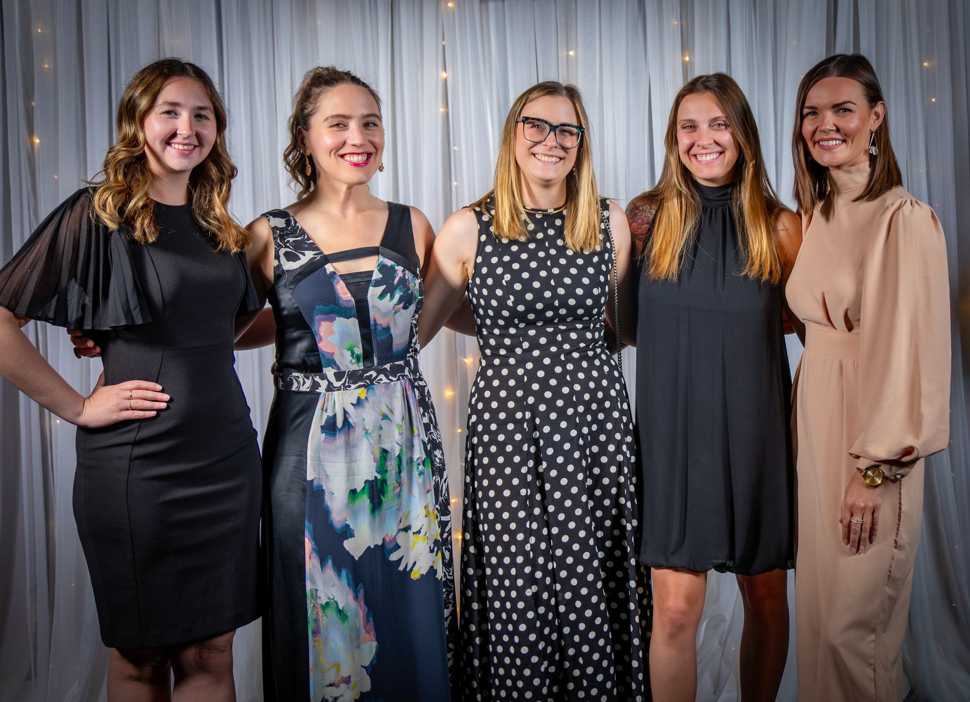 Five women smiling, arm-in-arm, posing in front of a white curtain with string lights.
