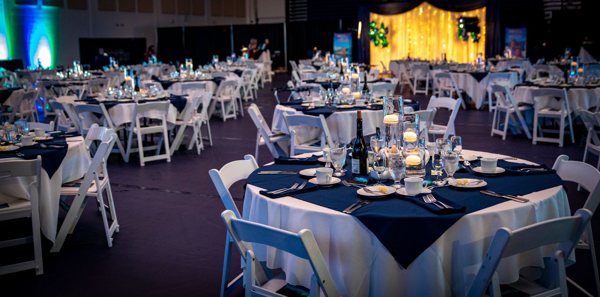 Elegant banquet hall with round tables set for an event, covered in white and navy linens, lit by candles.
