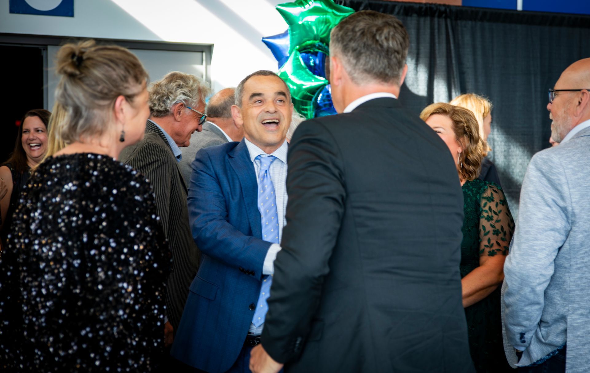 People mingling at a lively indoor event, with a man in a blue suit smiling in conversation.