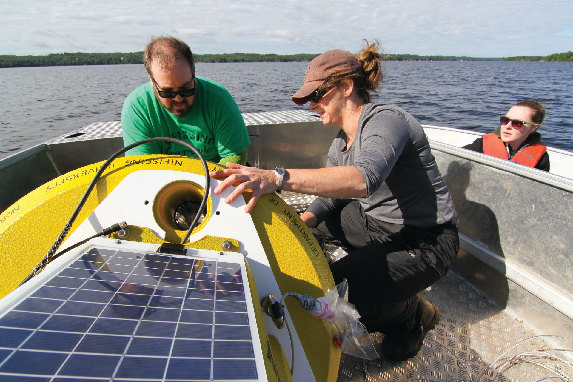 Three people on a boat tending a yellow buoy with a solar panel on a lake.