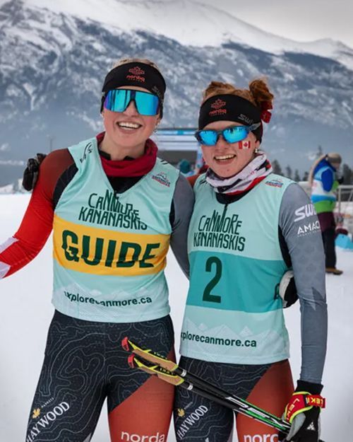Two skiers in teal race bibs and mirrored sunglasses standing on a snowy course with mountains behind them