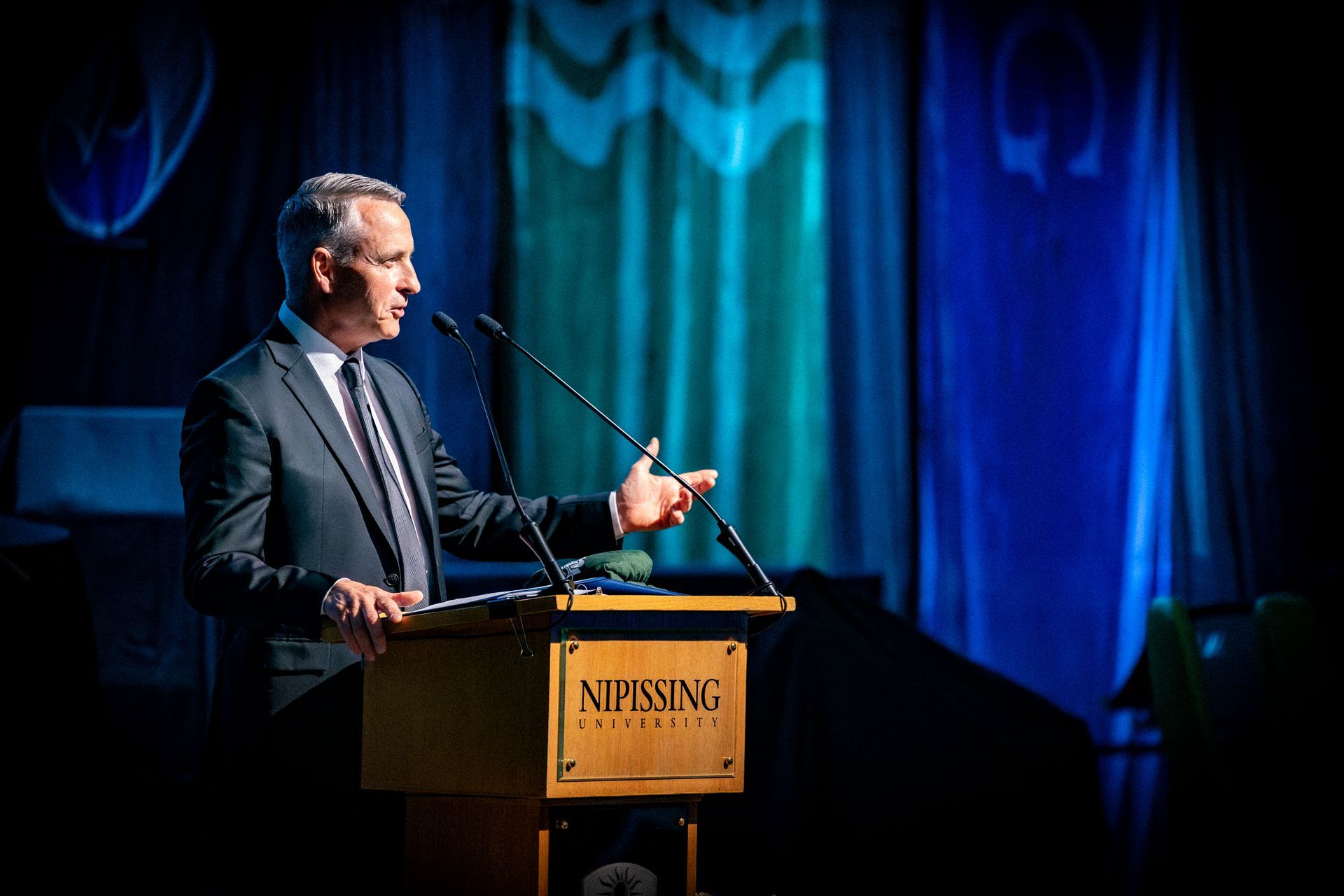 Black-and-white speaker at a podium, gesturing while addressing an audience on a dimly lit stage.